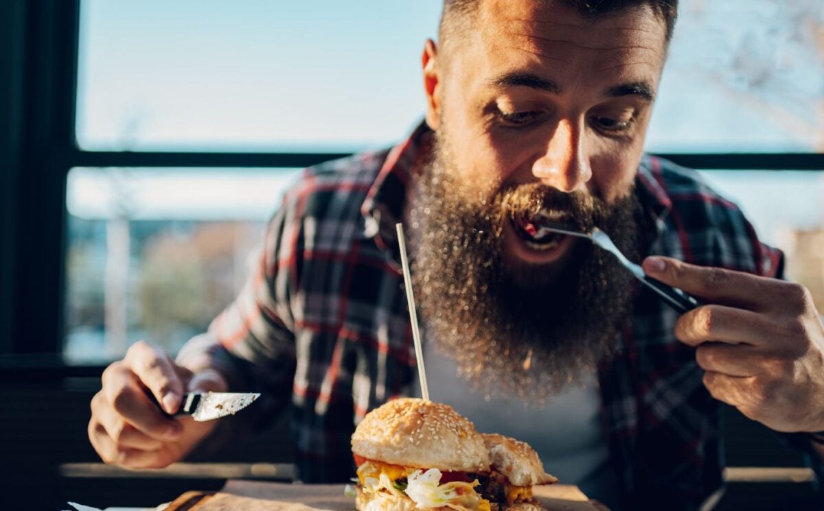 Photo shows a bearded man eating a burger with a knife and fork