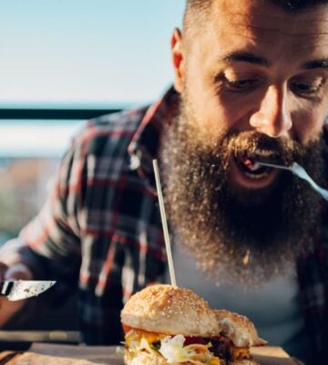 Photo shows a bearded man eating a burger with a knife and fork