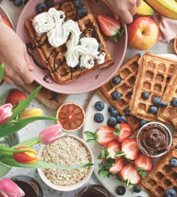 a spread of apple waffles served with fruit, vegan chocolate, and whipped cream