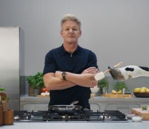 Gordon Ramsay stands in a grey kitchen while a cow hands him a spatula, part of a new campaign with Flora