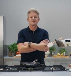 Gordon Ramsay stands in a grey kitchen while a cow hands him a spatula, part of a new campaign with Flora