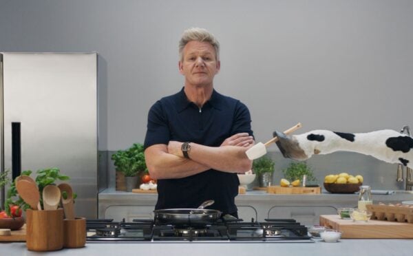 Gordon Ramsay stands in a grey kitchen while a cow hands him a spatula, part of a new campaign with Flora