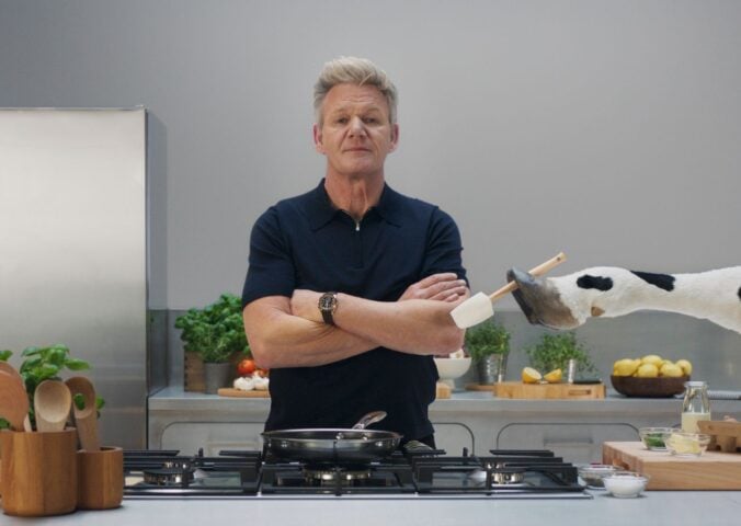 Gordon Ramsay stands in a grey kitchen while a cow hands him a spatula, part of a new campaign with Flora