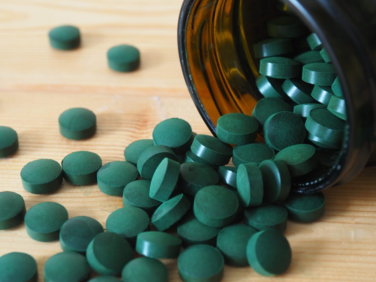 Photo shows a bottle of dark green supplement tablets spilling onto a wooden table