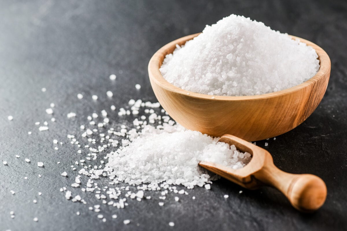 Photo shows a wooden bowl full of salt next to a wooden spoon on a grey surface