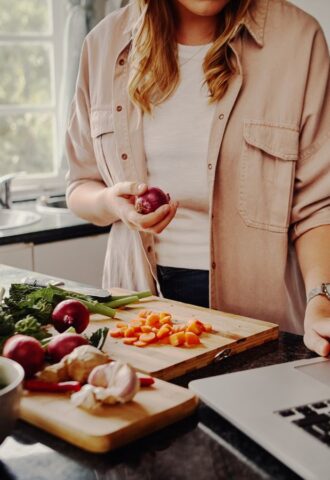 Photo shows a woman cooking fresh vegetables and using her laptop at the same time