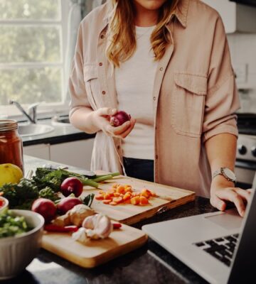 Photo shows a woman cooking fresh vegetables and using her laptop at the same time