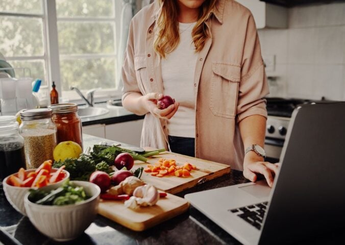 Photo shows a woman cooking fresh vegetables and using her laptop at the same time