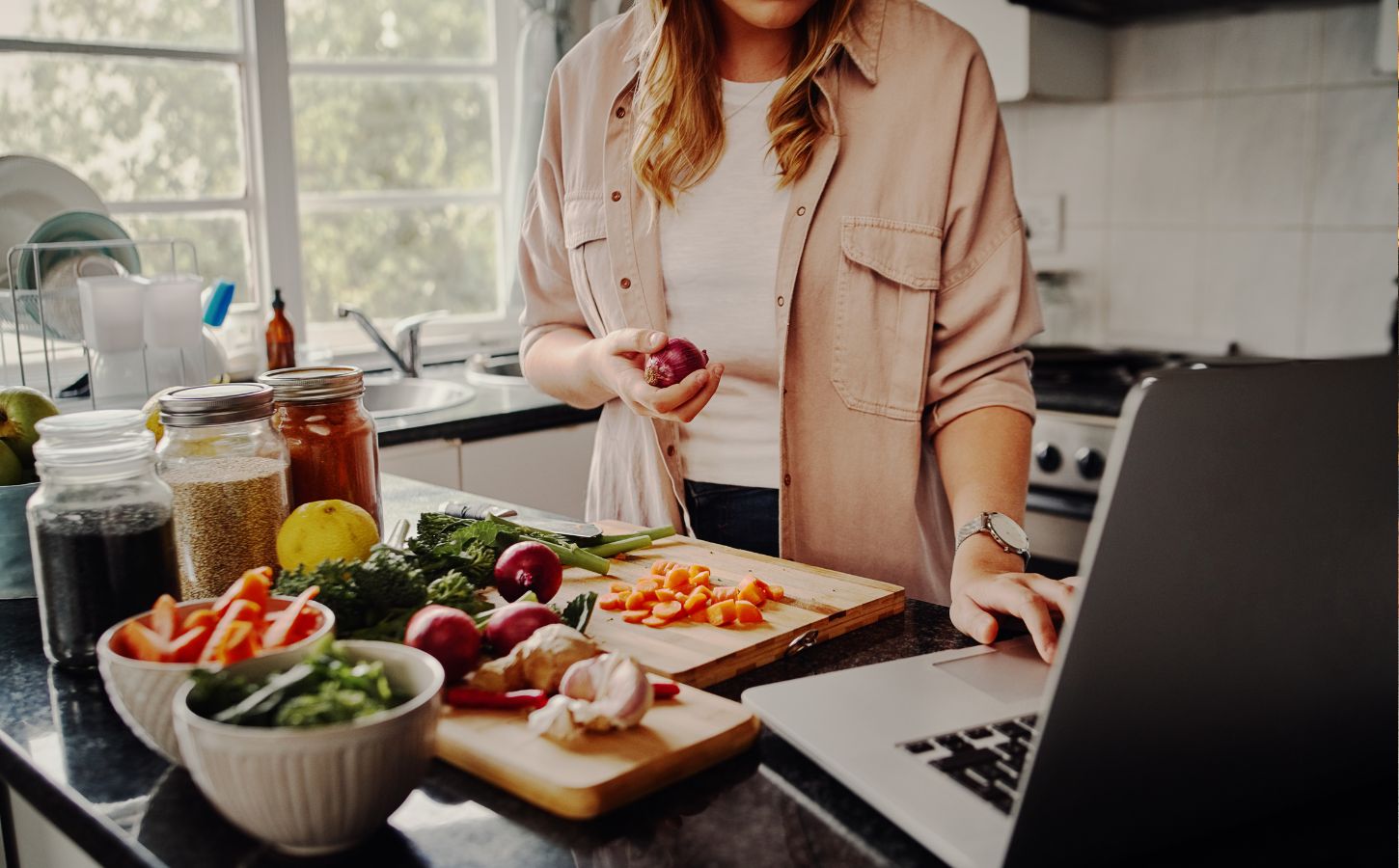 Photo shows a woman cooking fresh vegetables and using her laptop at the same time