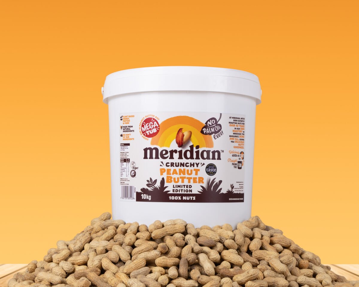 A large 10kg tub of peanut butter on top of a pile of peanut shells in front of an orange background