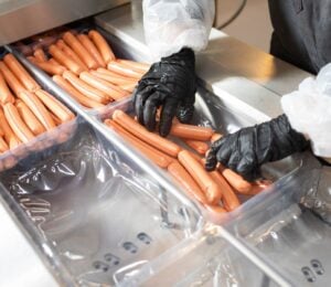 Photo shows a factory worker in a white coat and black rubber gloves placing many hotdogs into containers on a metal conveyor belt