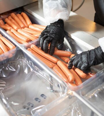 Photo shows a factory worker in a white coat and black rubber gloves placing many hotdogs into containers on a metal conveyor belt