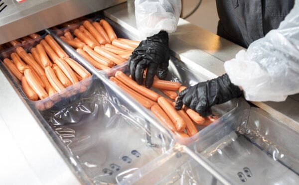 Photo shows a factory worker in a white coat and black rubber gloves placing many hotdogs into containers on a metal conveyor belt