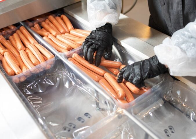 Photo shows a factory worker in a white coat and black rubber gloves placing many hotdogs into containers on a metal conveyor belt