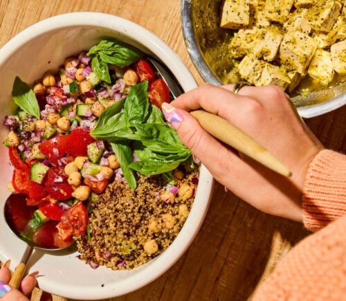 A quinoa bowl filled with chickpeas, tomatoes, cucumber, and basil, next to some dairy-free tofu feta