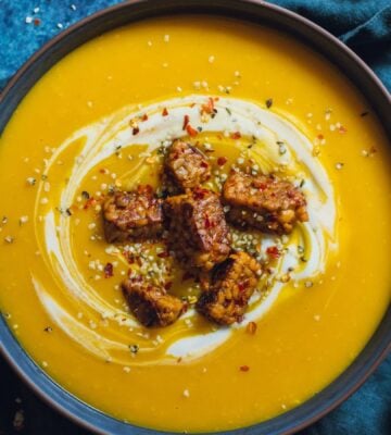 a bowl of spicy swede soup with ginger topped with vegan yogurt, chili, and tempeh chunks