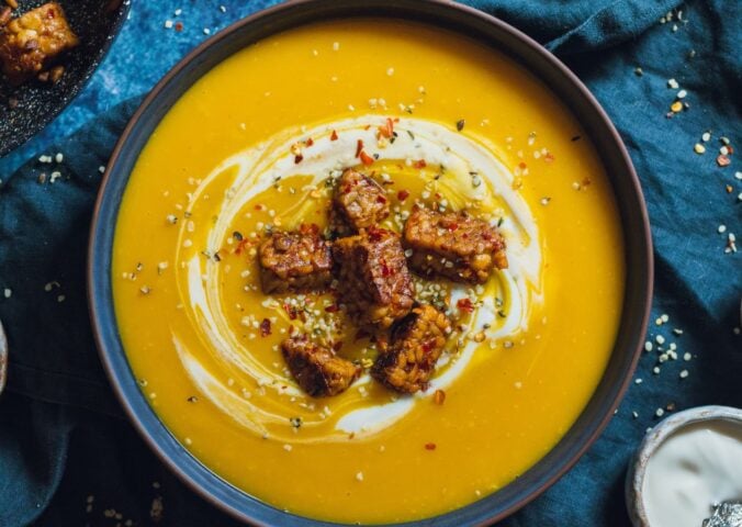 a bowl of spicy swede soup with ginger topped with vegan yogurt, chili, and tempeh chunks