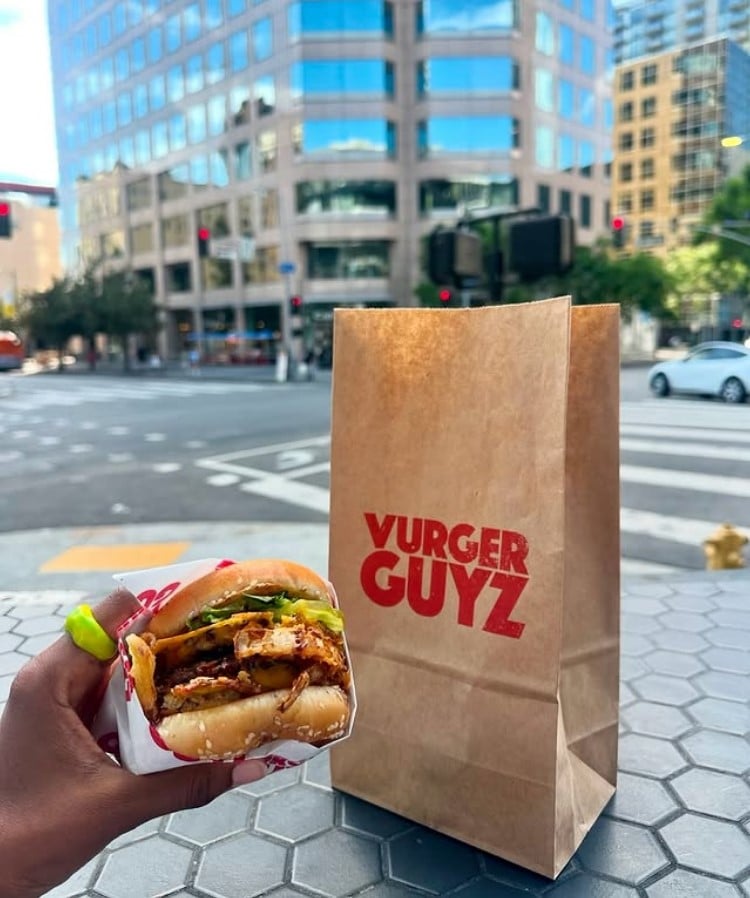 Photo shows someone's hand as they hold a burger up in front of a paper bag printed with the Vurger Guyz logo