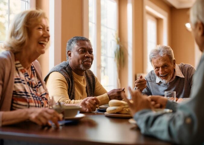 Photo shows a group of older people sat around a table eating and laughing together