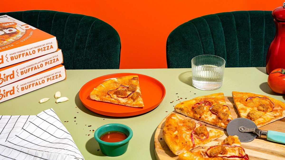 Photo shows several boxes of Blackbird Foods pizzas next to a plate and a pizza stone containing slices of cooked pizza