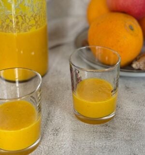 Two ginger and turmeric shots next to oranges and apples