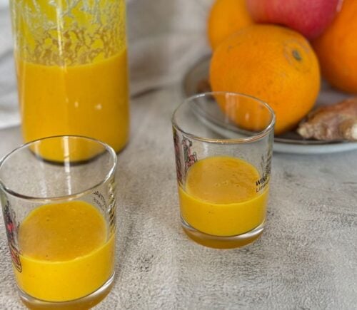 Two ginger and turmeric shots next to oranges and apples