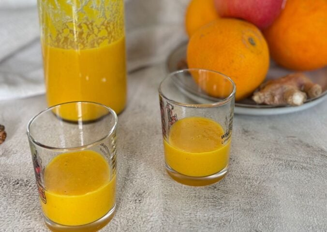 Two ginger and turmeric shots next to oranges and apples