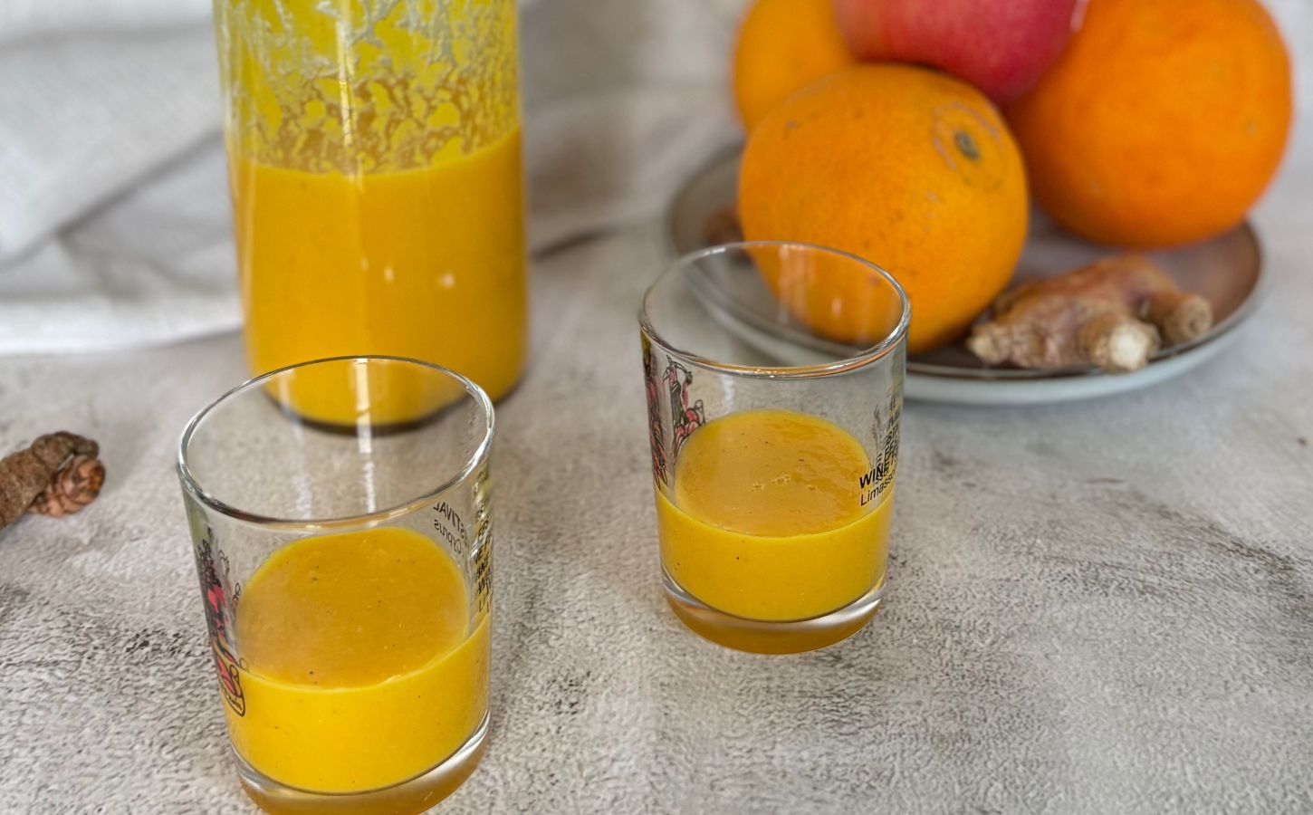 Two ginger and turmeric shots next to oranges and apples