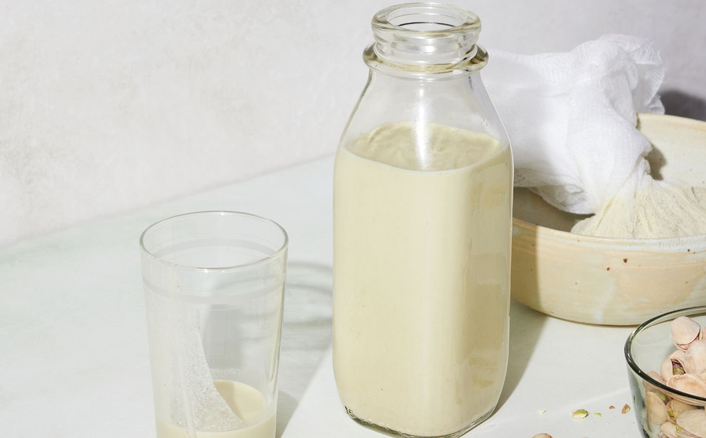 a picture of a bottle of perfect pistachio milk with s glass, cheesecloth, and whole pistachio nuts