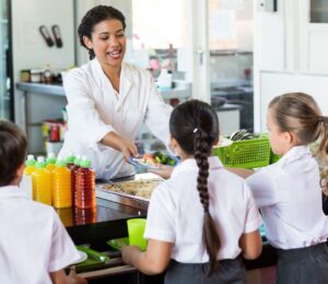 Photo shows an adult serving school children cooked vegetables in a cafeteria