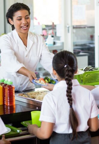 Photo shows an adult serving school children cooked vegetables in a cafeteria