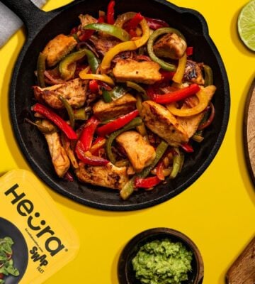 Photo shows a chopped up chicken fillet from Heura and Swap in a cast iron pan along with peppers and onions, against a yellow background, and alongside the product's new packaging