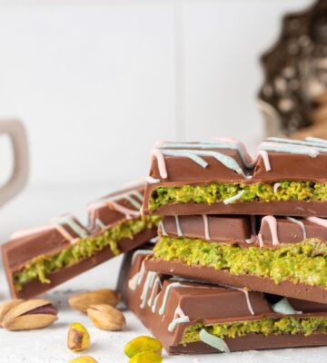 Photo shows a stack of Dubai Chocolate bar pieces, including a decorative pattern on the chocolate shell and a distinctive green filling, next to a cup of tea and pistachio shells