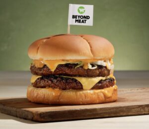 Photo shows a plant-based burger on a wooden board with a small Beyond Meat flag on a toothpick