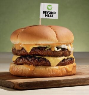 Photo shows a plant-based burger on a wooden board with a small Beyond Meat flag on a toothpick