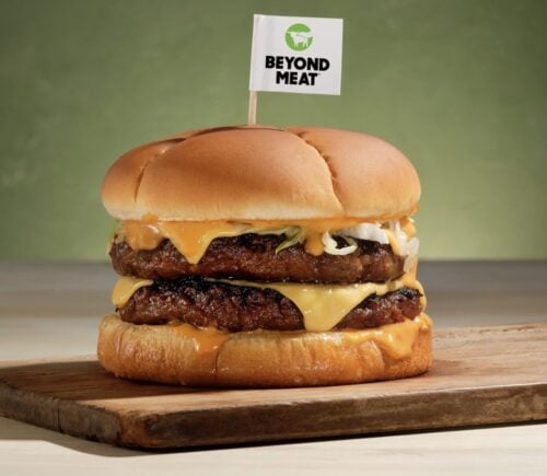 Photo shows a plant-based burger on a wooden board with a small Beyond Meat flag on a toothpick