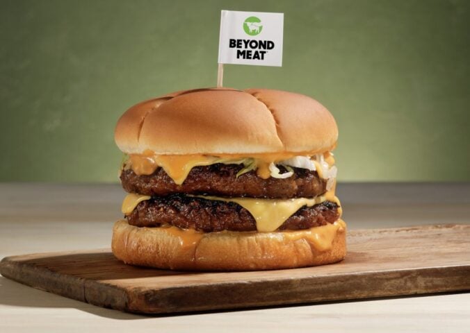 Photo shows a plant-based burger on a wooden board with a small Beyond Meat flag on a toothpick