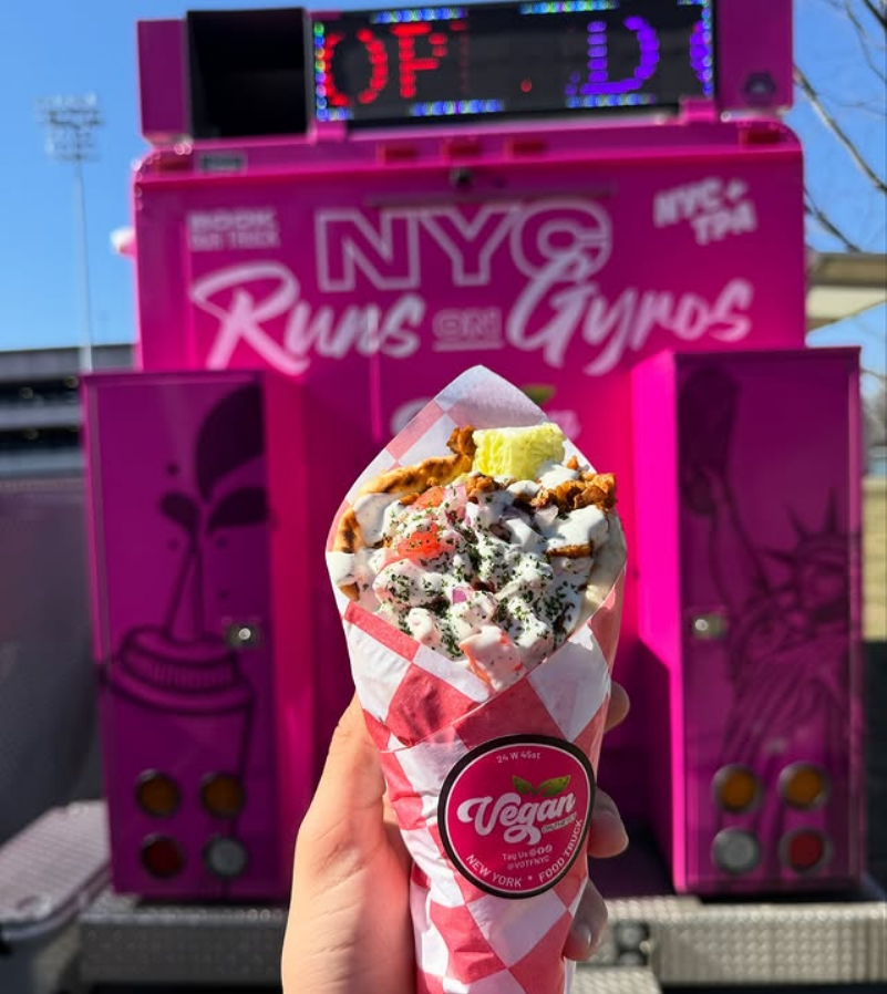 Photo shows someone's hand as they hold up a vegan gyro in front of a pink sign for Vegan On The Fly