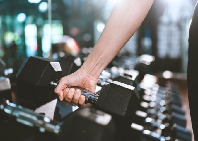Photo shows a woman's hand as she moves to pick up a dumbbell in a gym