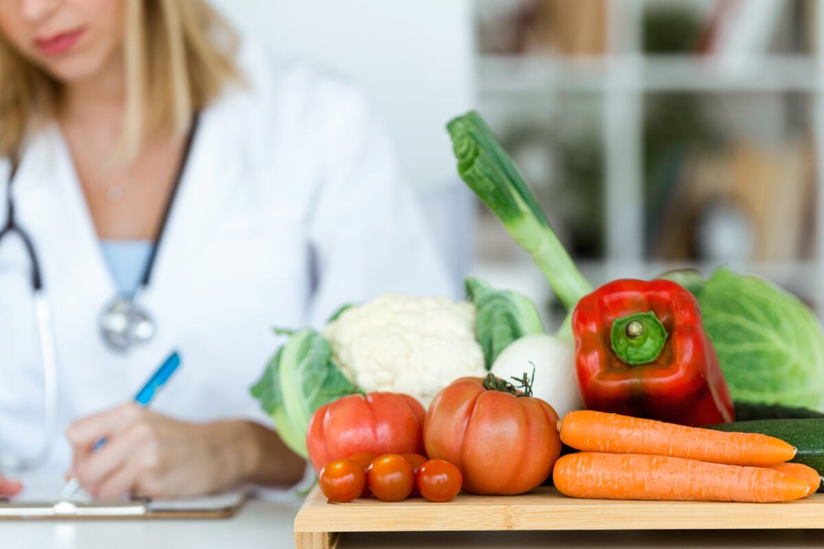 Photo shows a Dr in a white coat writing on a clipboard next to a chopping board and fresh vegetables
