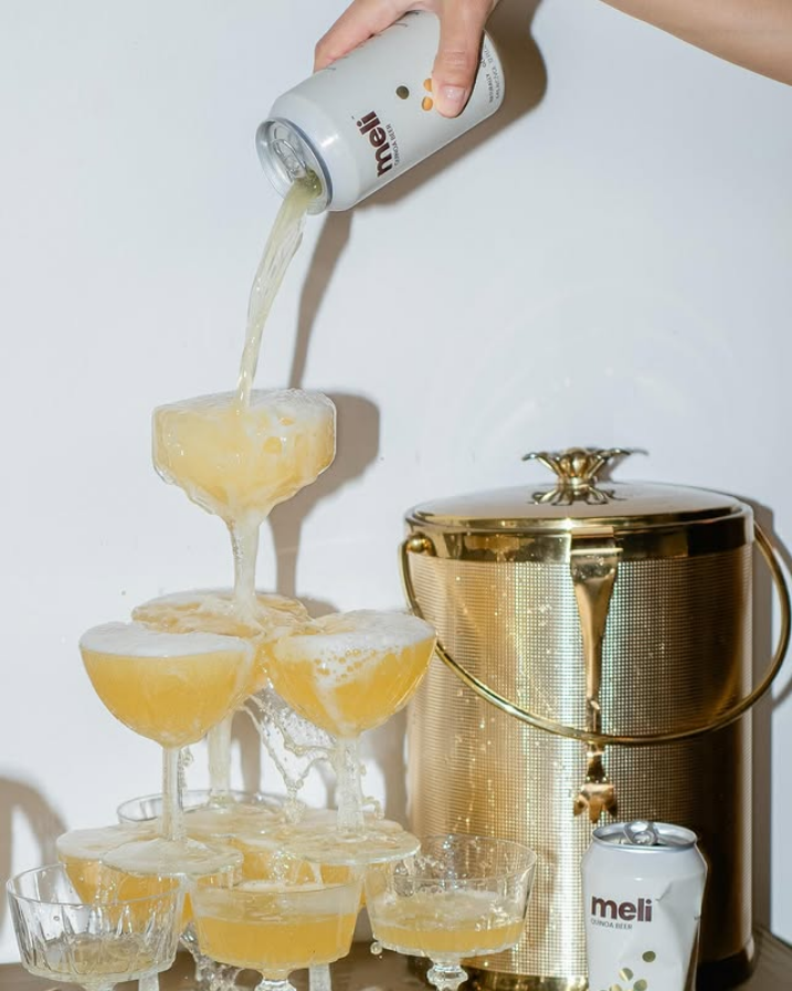 Photo shows someone pouring a can of Meli into overflowing champagne glasses