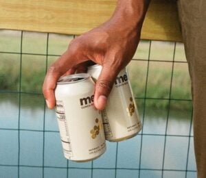 Photo shows someone's hand as they hold out two cans of Meli beer on a bridge above a river