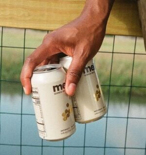 Photo shows someone's hand as they hold out two cans of Meli beer on a bridge above a river