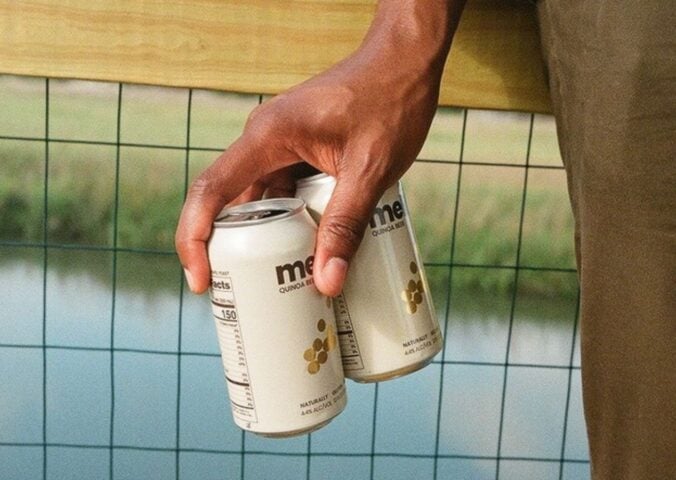 Photo shows someone's hand as they hold out two cans of Meli beer on a bridge above a river