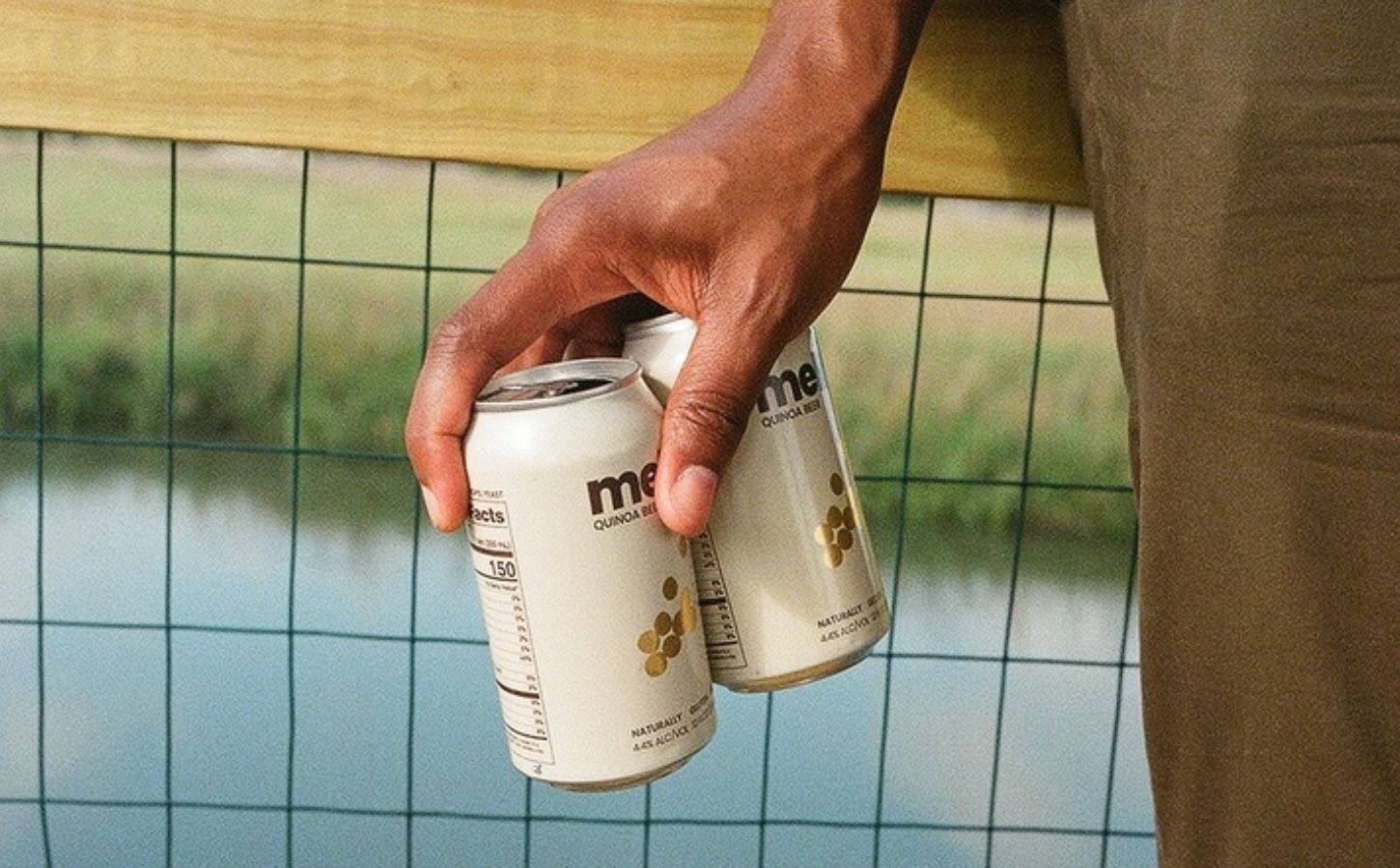 Photo shows someone's hand as they hold out two cans of Meli beer on a bridge above a river