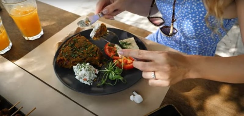 Charlotte, from Vegan Explores, is sitting down to eat a delicious-looking vegan meal in a tropical location on a wooden picnic table with a couple of juice glasses on it.