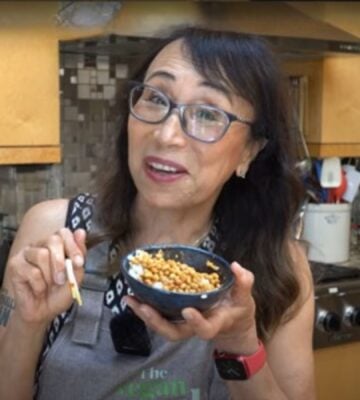 Woman wearing apron standing in kitchen, smiling and holding chopsticks and a bowl of white rice and natto.