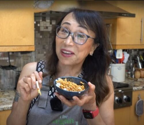 Woman wearing apron standing in kitchen, smiling and holding chopsticks and a bowl of white rice and natto.