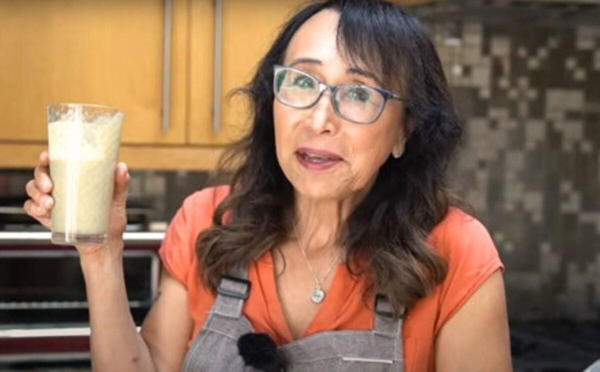 Woman with black hair and glasses holds up glass of protein shake as she stands in a kitchen.