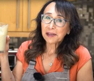 Woman with black hair and glasses holds up glass of protein shake as she stands in a kitchen.